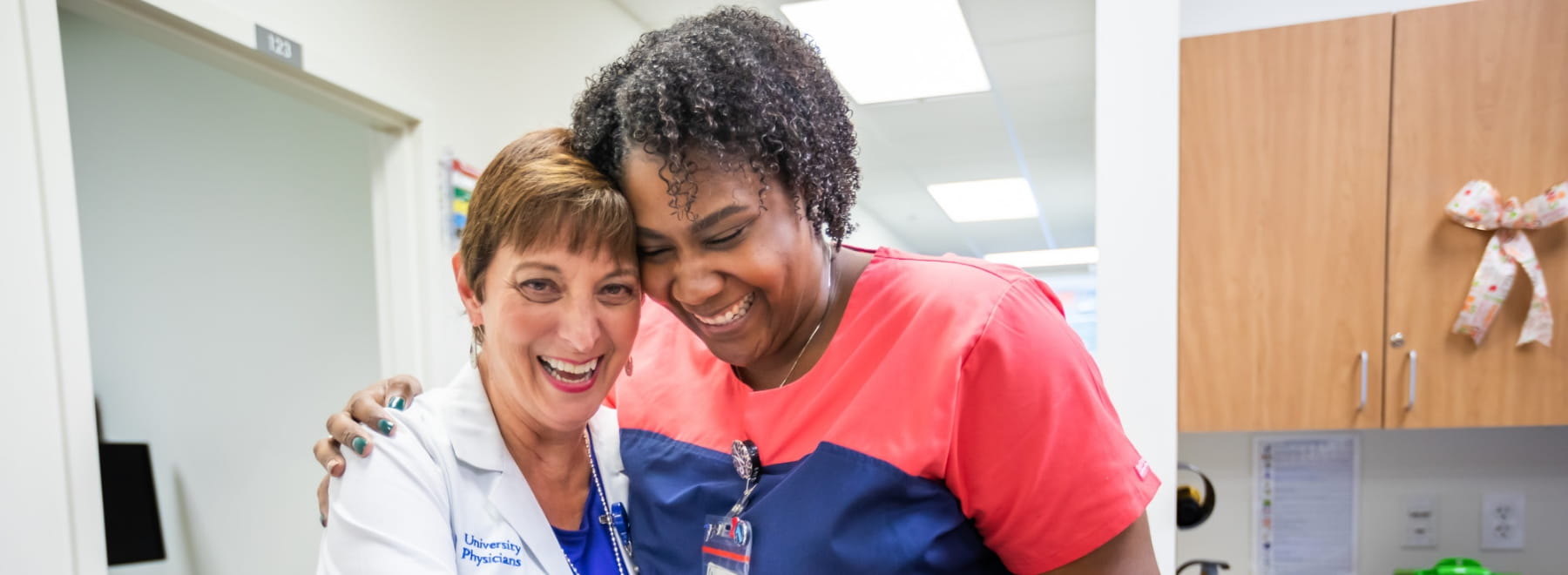 Female physician and female nurse smile while hugging next to a nurses station