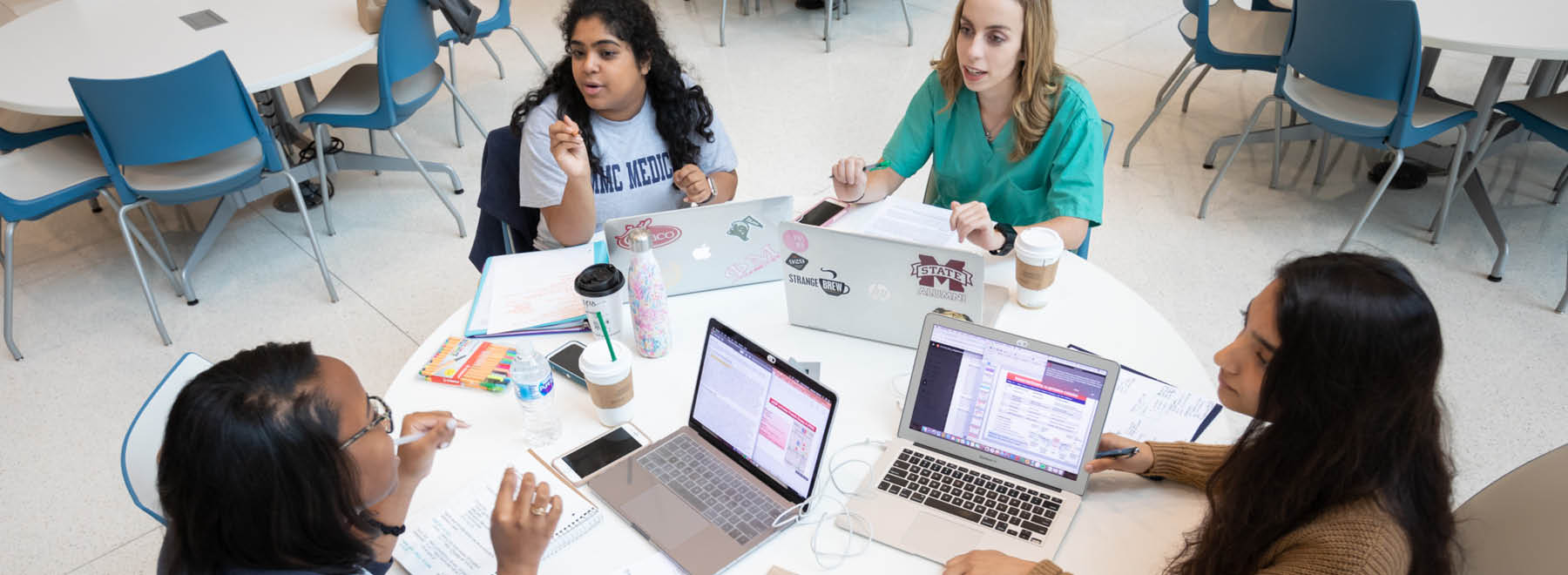 A group of five female medical students sit around a table, talking, with their laptops in front of them.