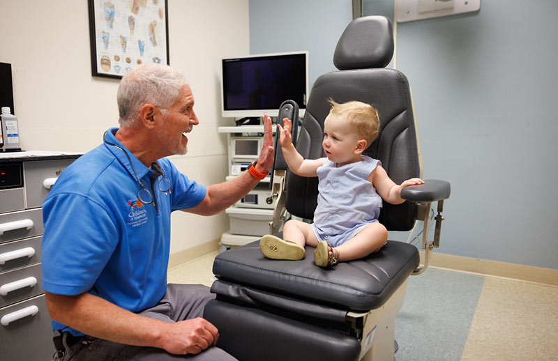 Pediatric visit. A pediatrician in a blue shirt smiles and gives a high-five to a toddler sitting on an exam chair in a medical office.