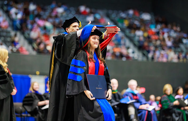 Graduation ceremony. A faculty member places an academic hood over a graduate’s shoulders during a university commencement ceremony in an arena.