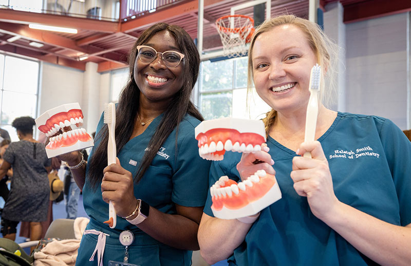 Dental students demonstration. Two dental students wearing teal scrubs hold oversized toothbrushes and large dental models while smiling for the camera.