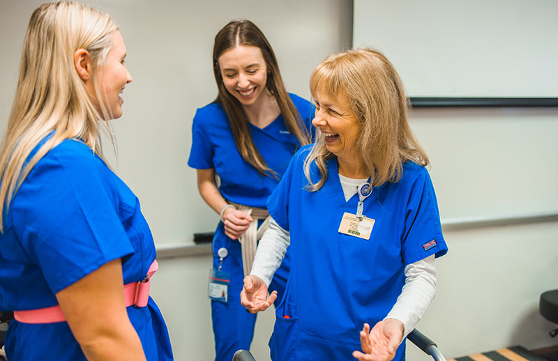 Nursing students laughing. Three people in blue scrubs laugh and talk together in a classroom or training room setting.
