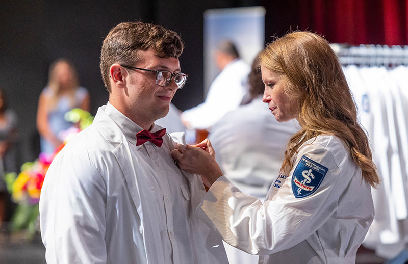 White coat ceremony. A faculty member helps a student adjust his white coat during a white coat ceremony, both smiling on stage under bright lights.