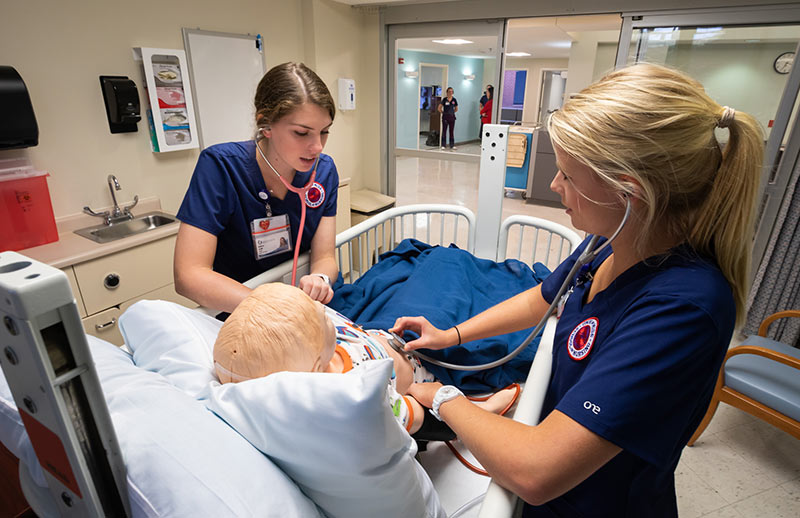 Nursing simulation. Two nursing students in navy scrubs practice using stethoscopes on a medical mannequin in a hospital simulation lab.