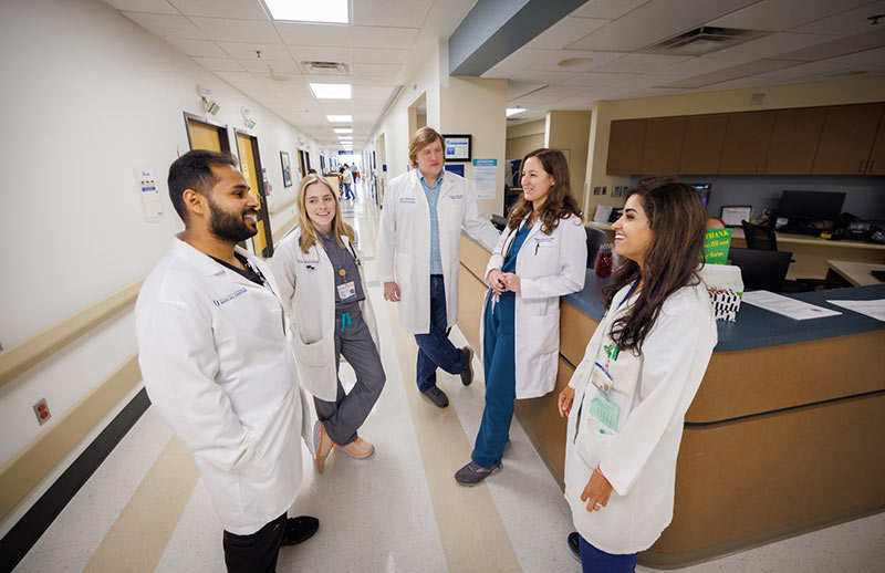 Group of clinicians in hallway. Five medical professionals wearing white coats stand together and talk in a hospital hallway near a nurses’ station.