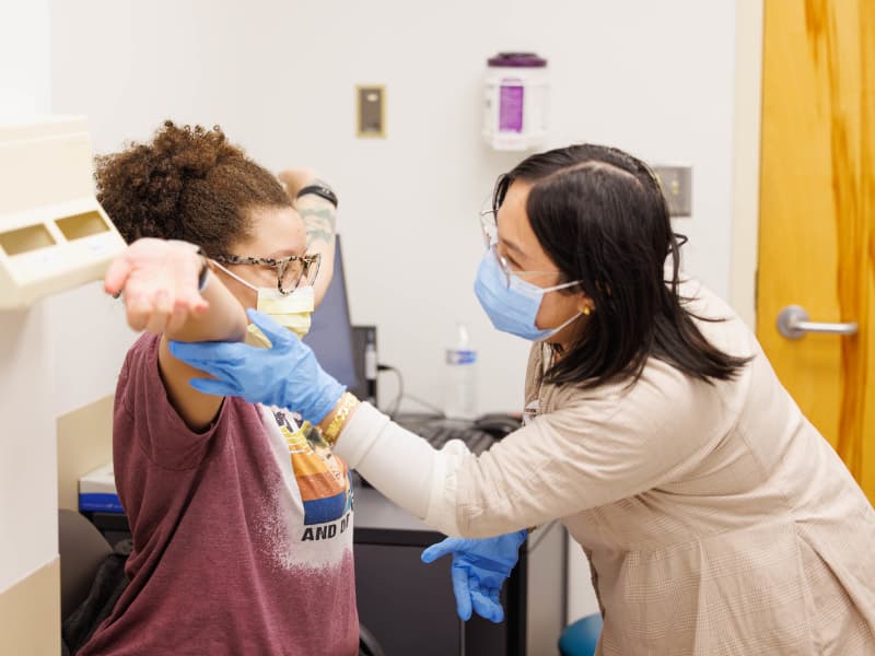 A patient sitting in a genetic clinic sits with her arms outstretched as a health professional examines her.