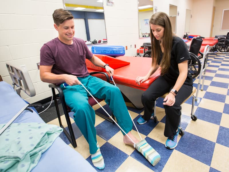 Audrey Hartman, a third year OT student, teaches classmate and simulated patient, Charles Nosco, how to put on his socks with the use of adaptive device. Nosco is simulating a patient who has had a hip replacement and is not allowed to bend over or pull knees up toward chest.