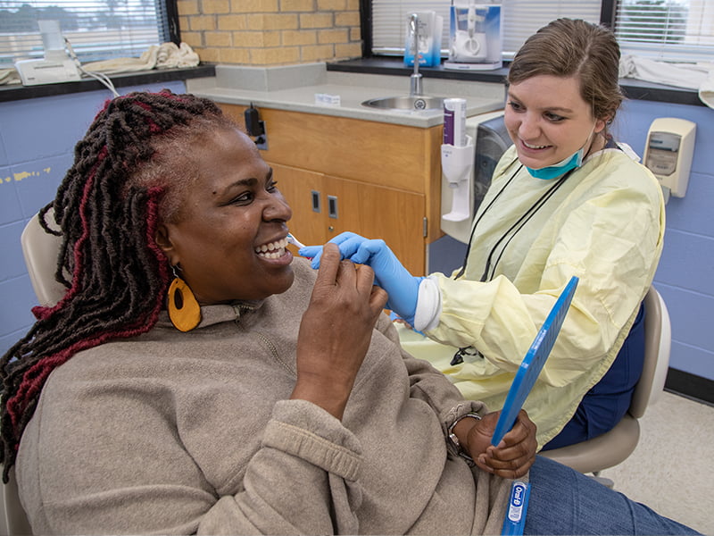 Woman in dental chair smiles as dental assistant shows toothbrush use.