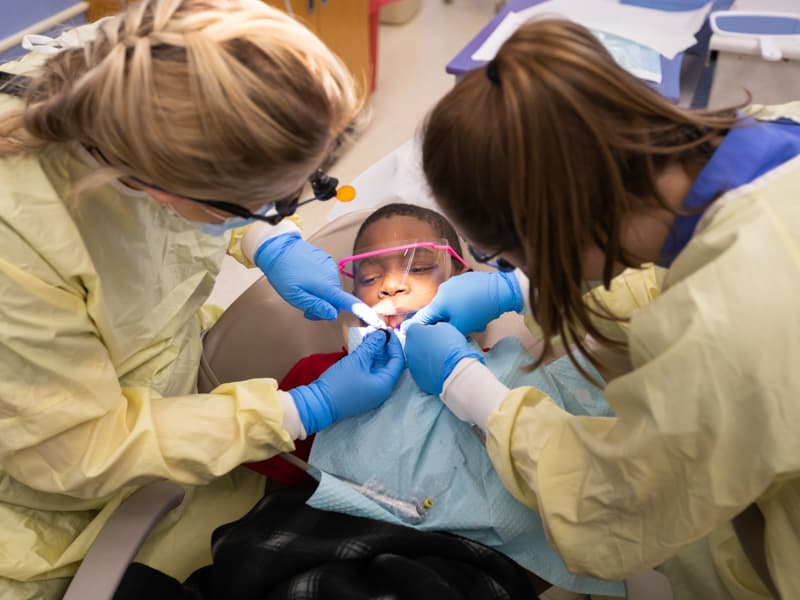 Two dental students lean over a pediatric patient while working.