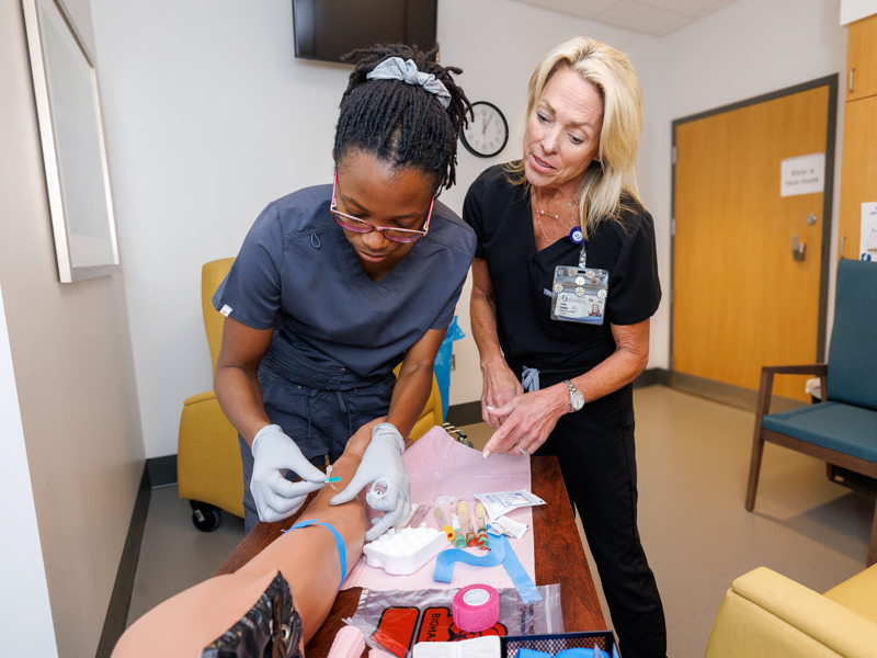 Cathy Hudgins, right, nursing manager for clinical reseach, teaches phlebotomy techniques to Rachael Duffin, research specialist, using a simulation arm. Duffin is an enrollee in the Clinical Research Coordinator Certificate Program. Hudgins is her preceptor.