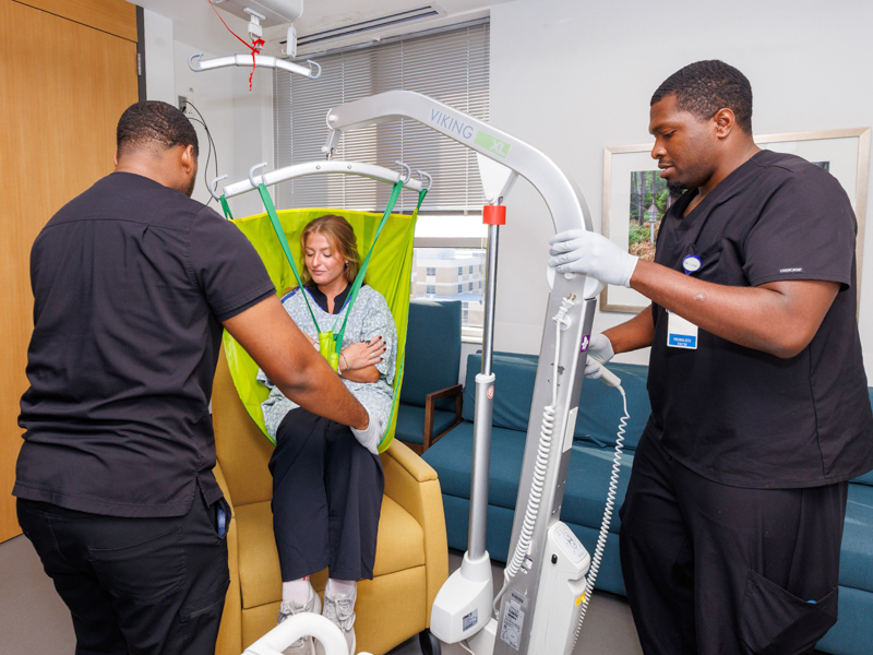 Mobility technicians Tavian Johnson, left, and Amos Hightower work with new technician Mary Addison McDaniel to train her to lift patients from bed to bedside chair.
