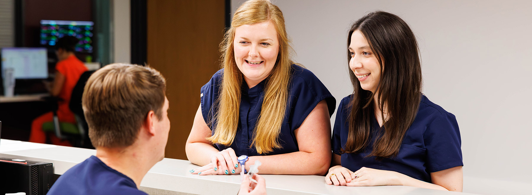 Three pharmacy residents chat at a workstation. One is seated and holding a syringe.