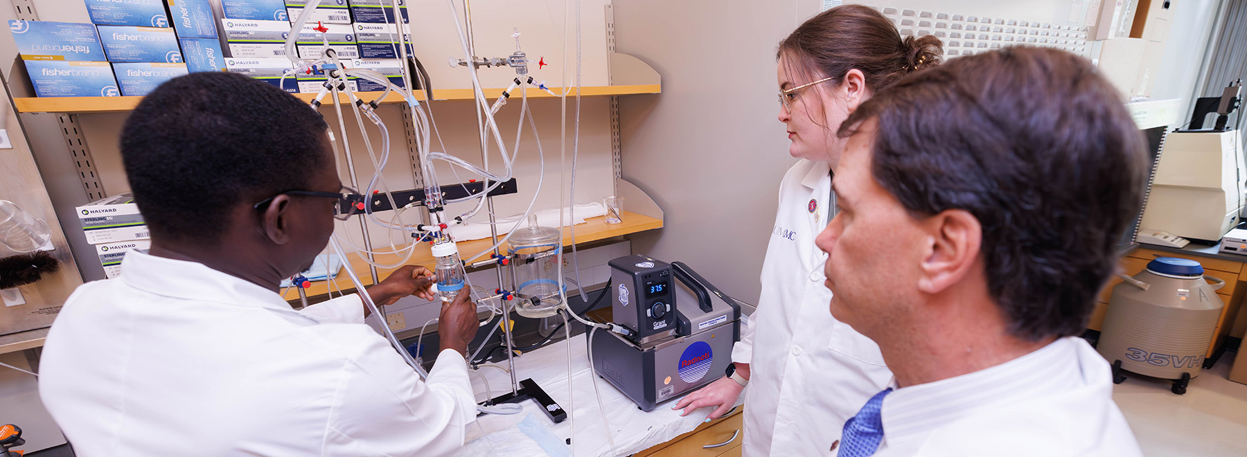 Dr. David Stec and a researcher, right, look on as another researcher examines a laboratory setup with tubing, glass containers, and scientific equipment on a workbench.