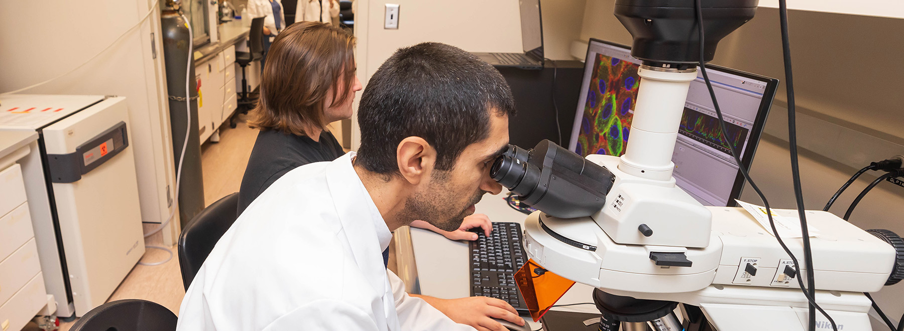 One researcher looks through a microscope as another in the background reviews data on a computer monitor.