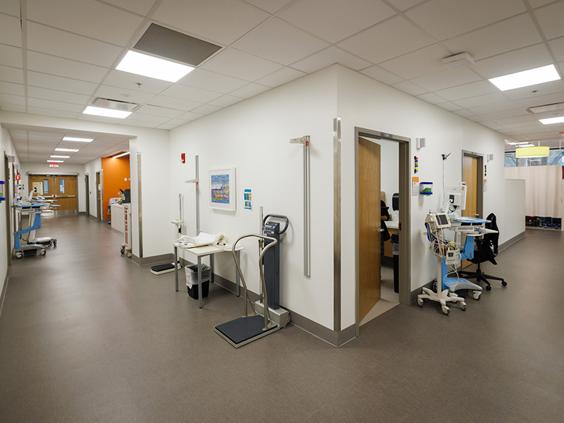 Healthcare clinic corridor with exam rooms, medical equipment workstations, patient scale, open staff office, and treatment areas extending from a central hallway.