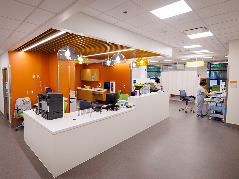 Open clinical nursing station with white reception counters, orange accent walls, pendant lighting, work areas, and curtained patient treatment bays in a modern outpatient clinic.