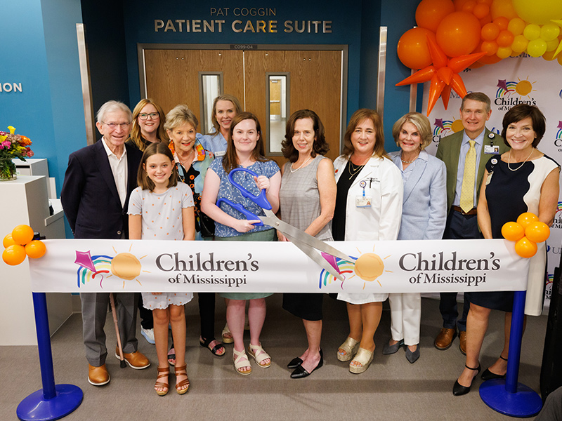 Ribbon-cutting ceremony at Children’s of Mississippi’s Pat Coggin Patient Care Suite, with a group gathered behind a branded ribbon and oversized ceremonial scissors beneath balloon decorations.