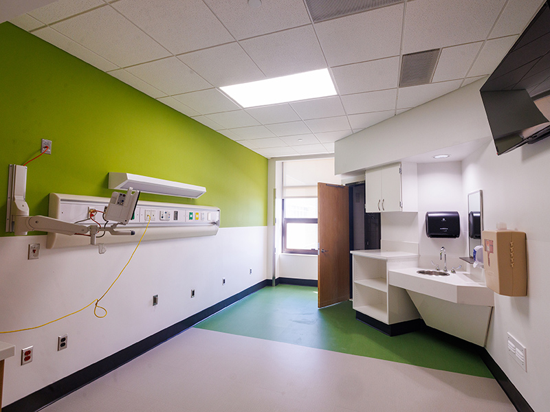 Patient room with medical gas and utility panel, green accent wall, wall-mounted monitor support, sink, and open doorway to adjoining clinical space.