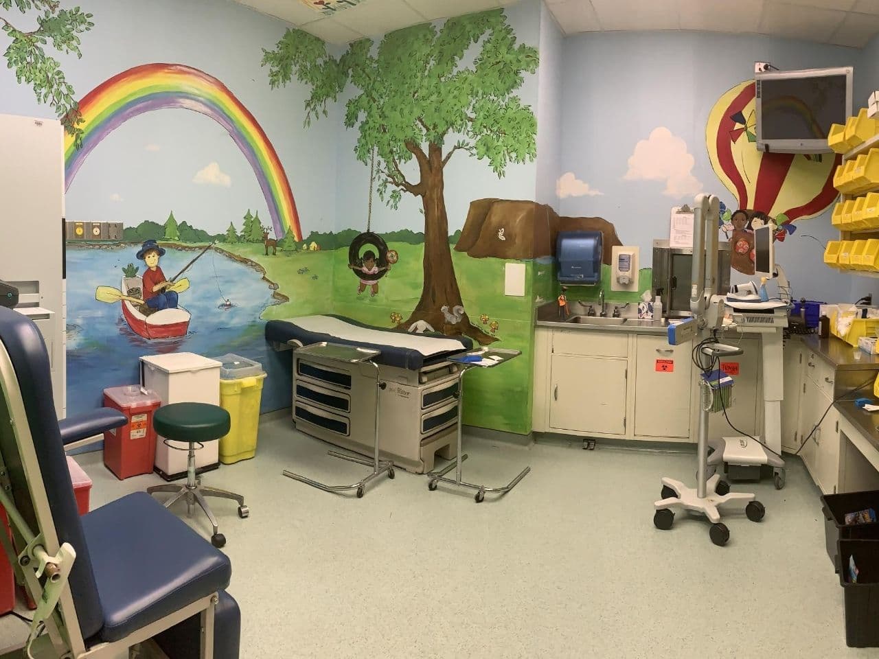 A procedure room has an exam table, chairs, counter space with sink, storage bins and is decorated with a full size wall mural showing a man fishing under a rainbow and a tree with a tire swing.