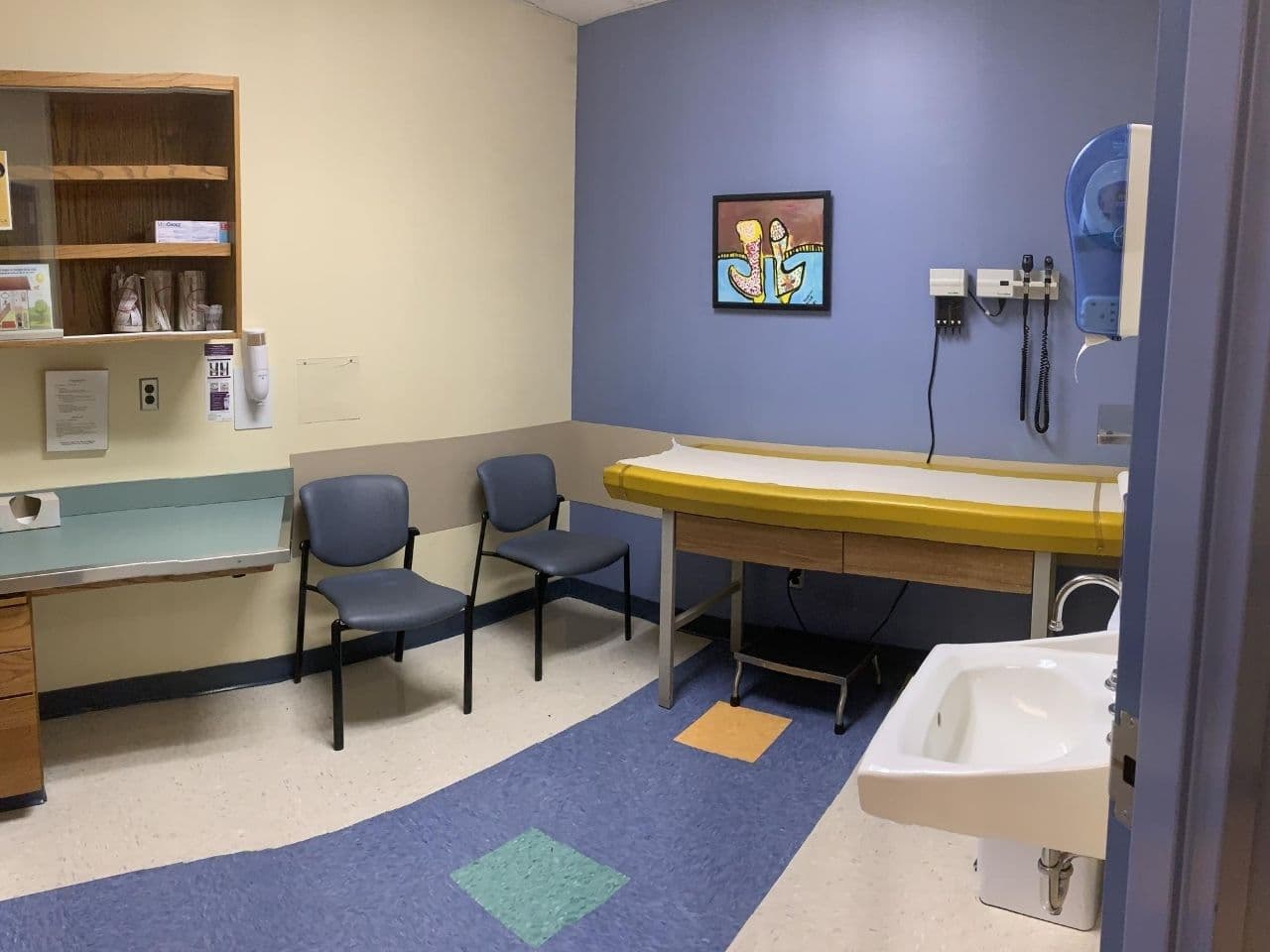 The exam room features a purple accent wall and a colorful painting along with exam table, sink, cabinets, and chairs.