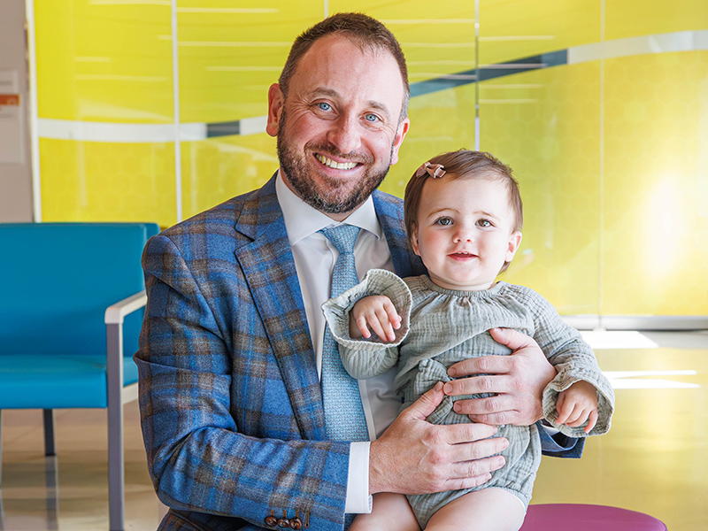 A seated Dr. Hoppe happily holds an infant ear reconstruction patient at the Morris Center within Sanderson Tower.