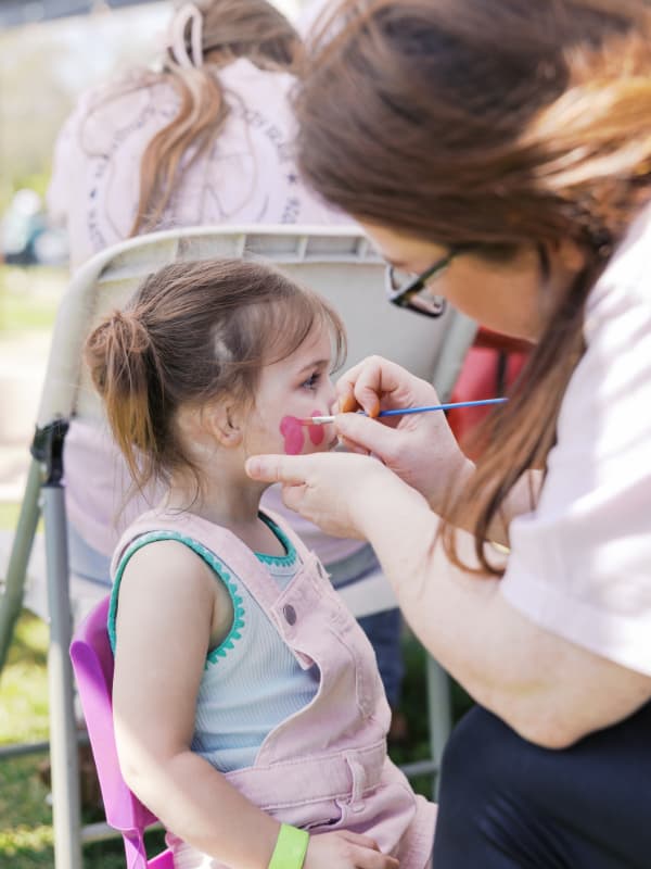 A girl in pink overalls sits  while a volunteer paints a butterfly on her face at Family Fun Day 2026.
