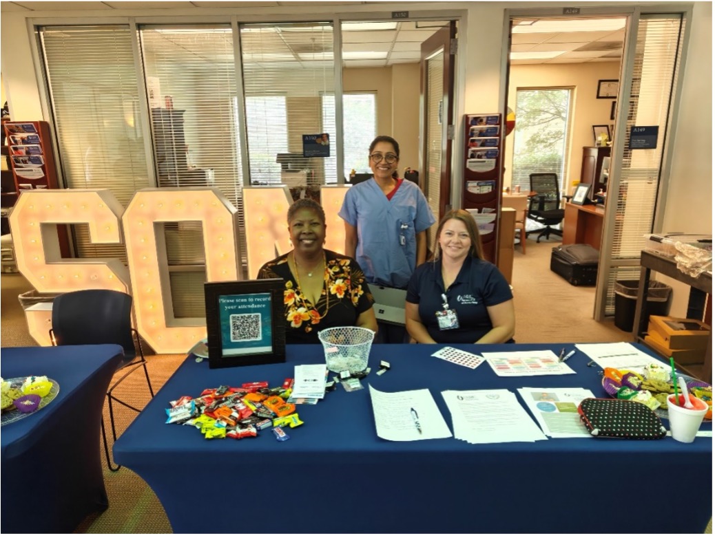 Three staff members sit and stand behind an information table covered with a blue tablecloth and outreach materials, including a QR code sign, handouts, and candy. Large illuminated “50M” letters are displayed behind the table inside an office suite with glass-walled offices and brochure racks.