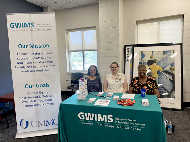 Information table for the GWIMS (Group on Women in Medicine and Science) at the University of Mississippi Medical Center. Three people are seated behind a teal GWIMS-branded table displaying brochures, candy, and giveaway items. A large standing banner beside the table lists the organization's mission and goals, including advancing participation and inclusion of women in academic medicine, promoting gender equity, recruitment and retention, awards and recognition, and career advancement. The table is set indoors near windows, with framed artwork displayed behind the group.