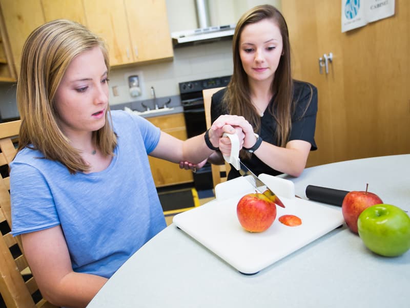 Second-year OT students, Mary Morgan Prince, left as simulated patient, and Anna Herrington, demonstrate how a patient with the use of only one upper extremity can learn to use adaptive equipment in the kitchen.