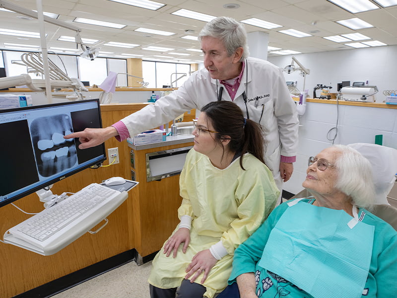 Dentist points at computer while dental student and patient look at the screen.