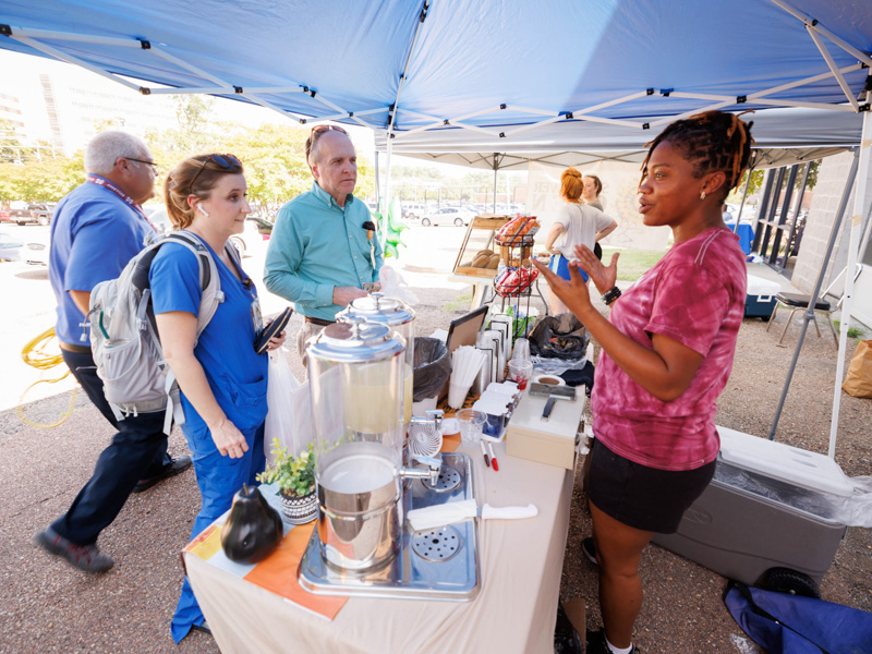 Keyah Williams with Mama Nature's Juice Bar in Fondren describes the bar's menu items to occupational therapist Emily Pierce, left, and DIS IT Project Manager Monte Luehlfing at Thursday afternoon's Farmer's Market behind Backyard Burger. The event was sponsored by the UMMC Office of Well-being. Joe Ellis/ UMMC Communications 