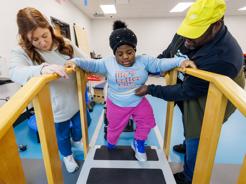 Makayla Campbell of Jackson, with support from physical therapist Kara Hudson and dad Maris Campbell, practices climbing stairs in the new Children's Rehabilitative Services space.