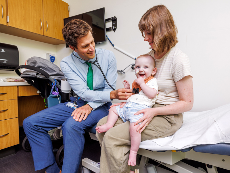 Dr. Erik Frandsen, left, examines a smiling baby with a stethoscope while the baby’s mother holds the child.