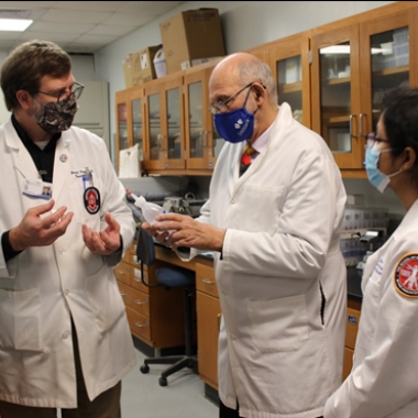 Two men and one woman wearing white coats and masks (Dr. Griggs, Dr. Mecholsky, and Megha Satpathy) discuss research progress.