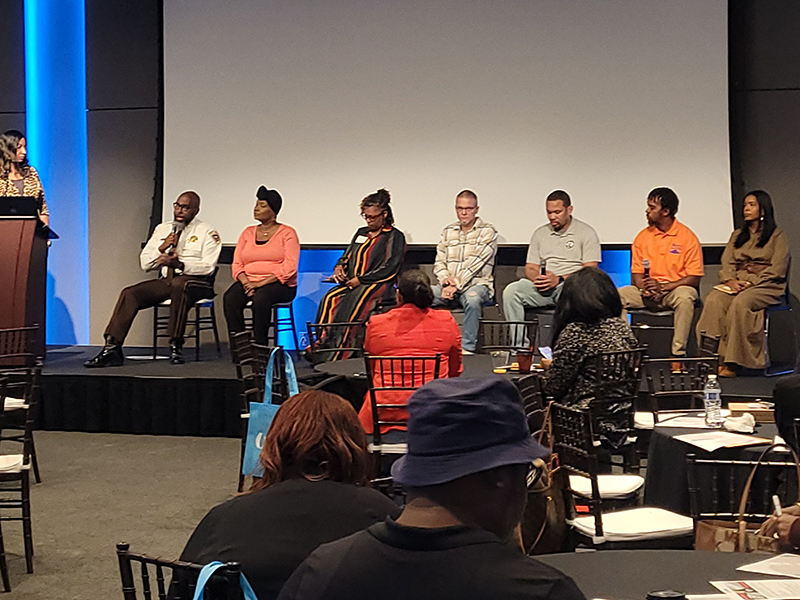 Seven panelists sitting on a stage as the first panelist, Sheriff Tyree Jones, addresses the crowd.