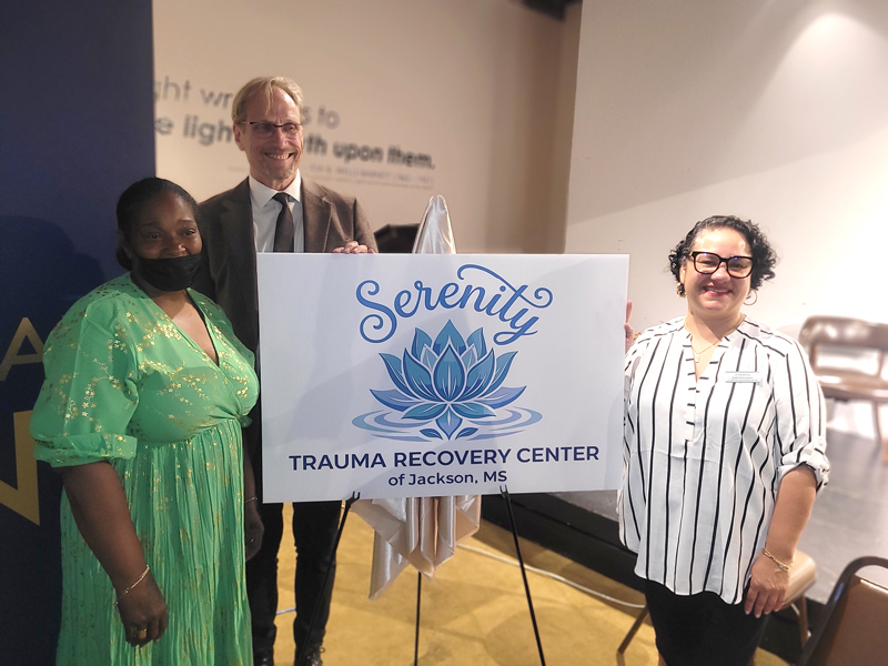 An event attendee, Rev. Dr. Martin Todd Allen and Sharon Brown stand around the sign with the Serenity Trauma Recovery Center logo.