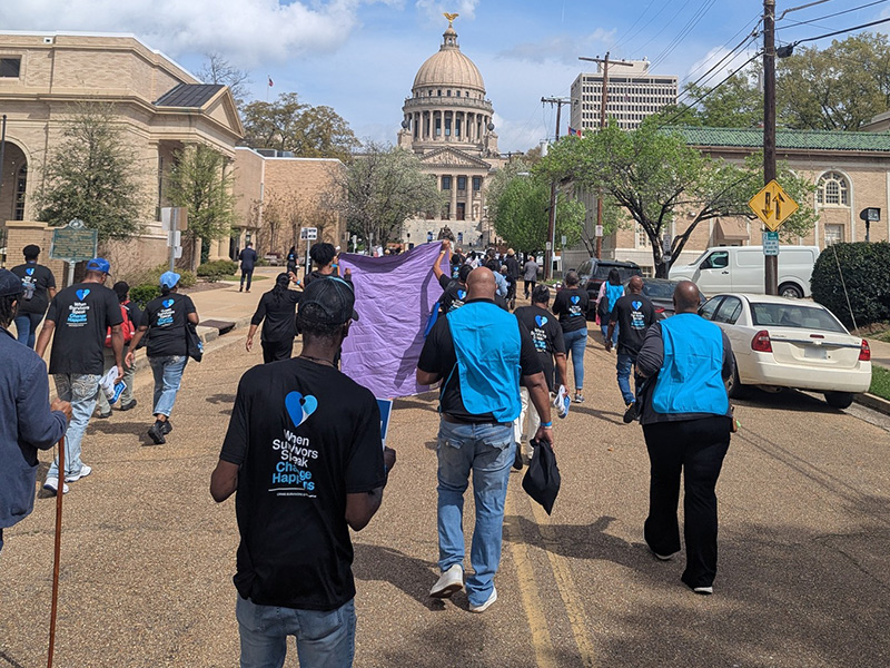 Group walking down a city street toward the Mississippi State Capitol carrying a purple banner during an organized march.
