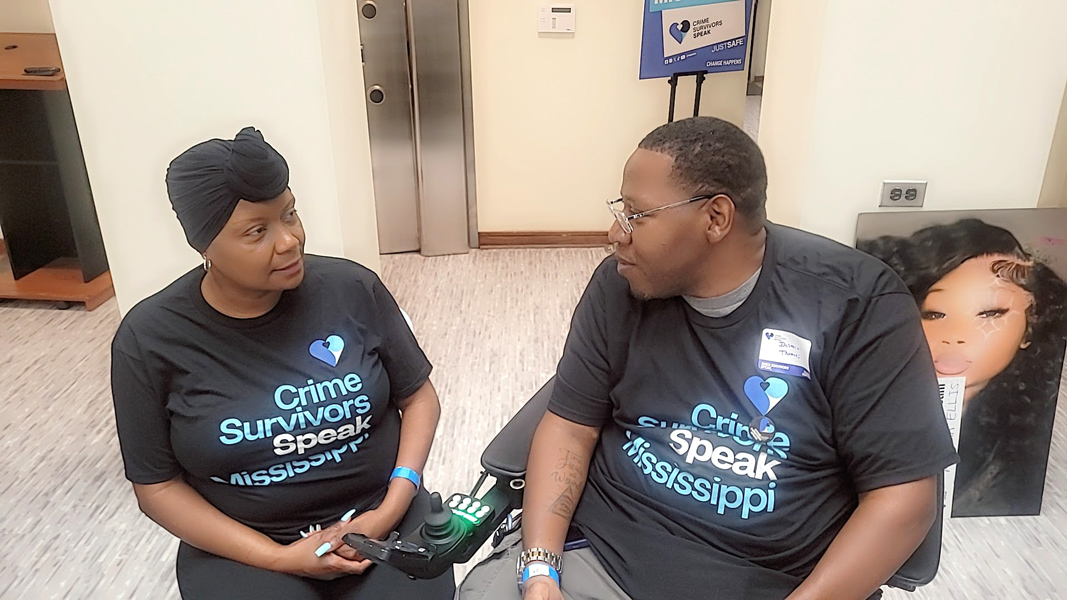 Two participants seated indoors looking at each other and wearing Crime Survivors Speak Mississippi shirts; one sits in a motorized scooter.