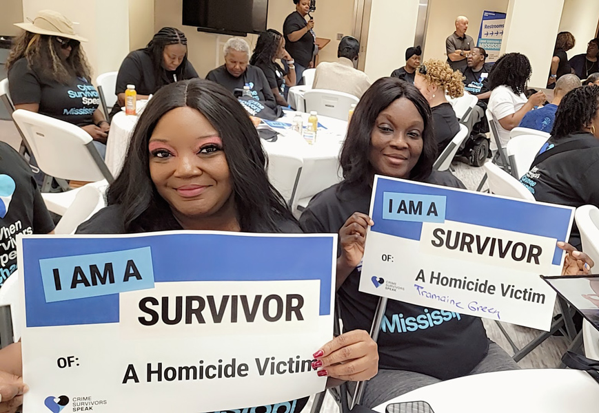 Two event attendees seated and holding signs that read I Am a Survivor of a Homicide Victim. The second sign has the name Tramaine Green.