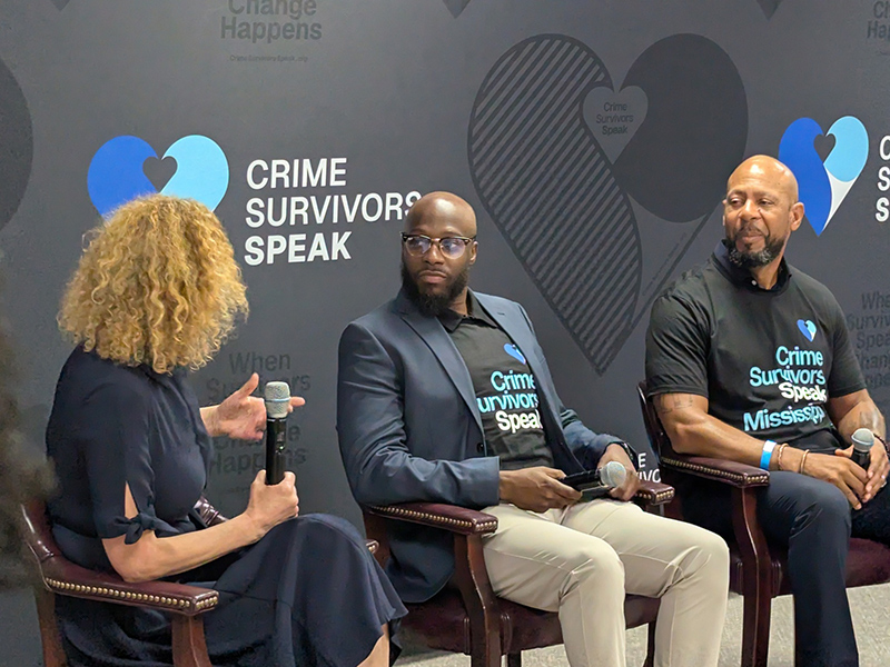 Panel discussion with three speakers seated on a stage in front of a Crime Survivors Speak backdrop, one holding a microphone.