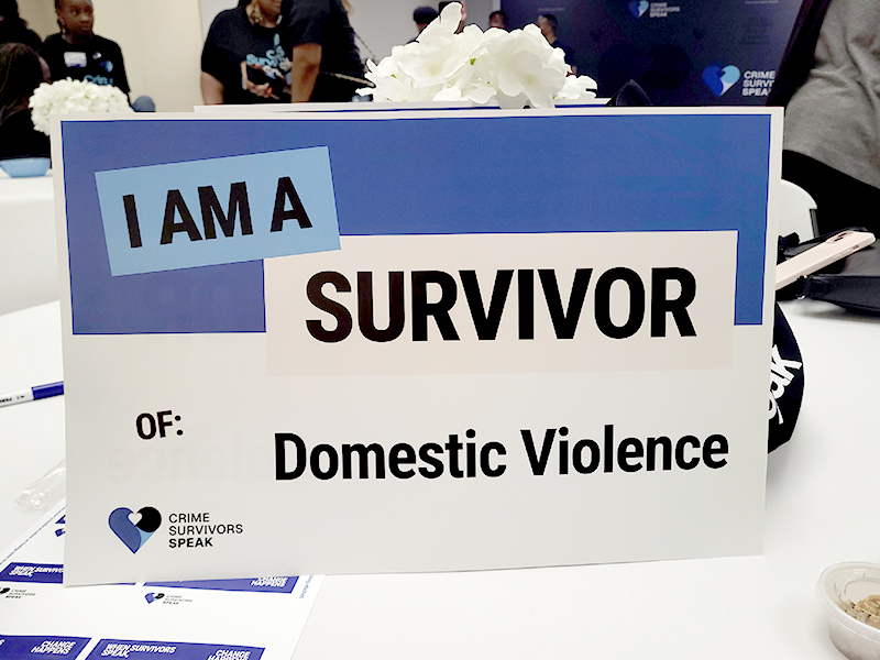 Table sign reading I Am a Survivor of Domestic Violence with people seated in the background.