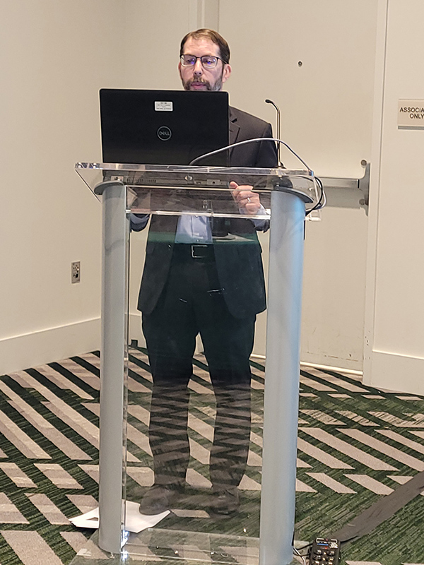 Conference presenter standing behind a clear podium and laptop while delivering a presentation in a meeting room.