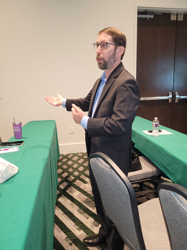 Person in business attire speaking during a conference session, standing between meeting tables and gesturing while addressing attendees in a training room.