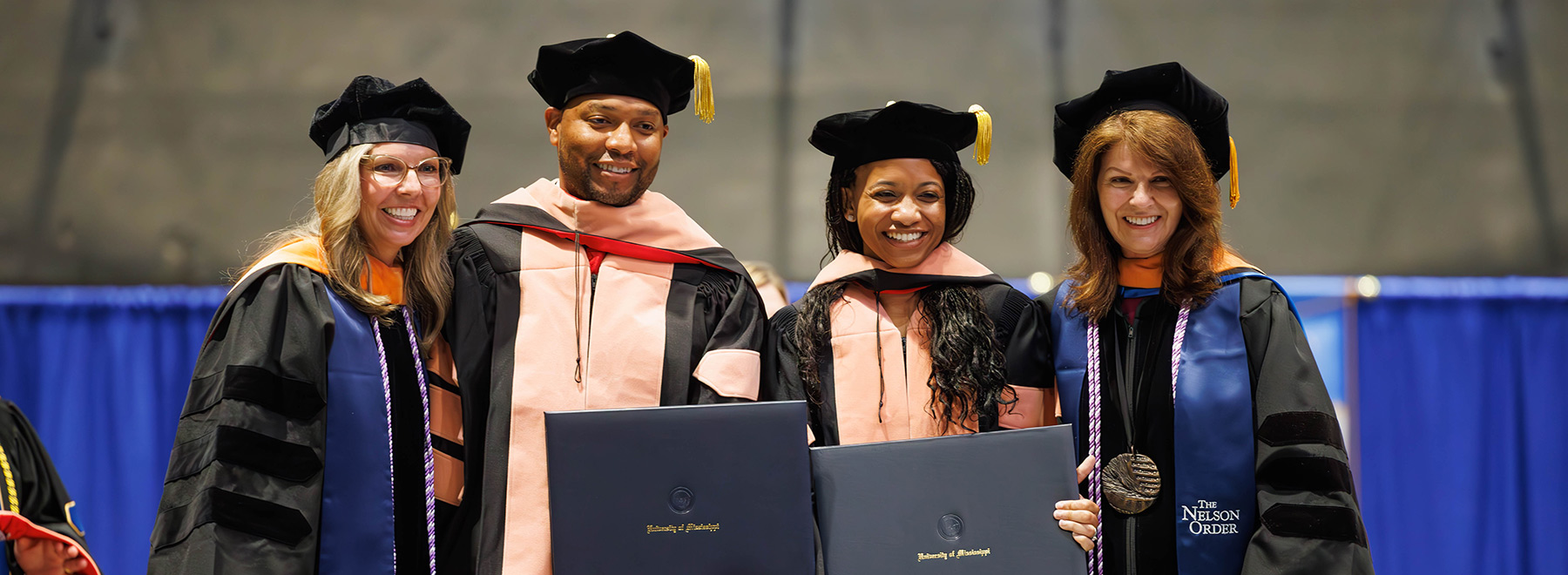 Graduates in academic regalia stand together holding diplomas during a commencement ceremony, wearing gowns, hoods, and caps with tassels.